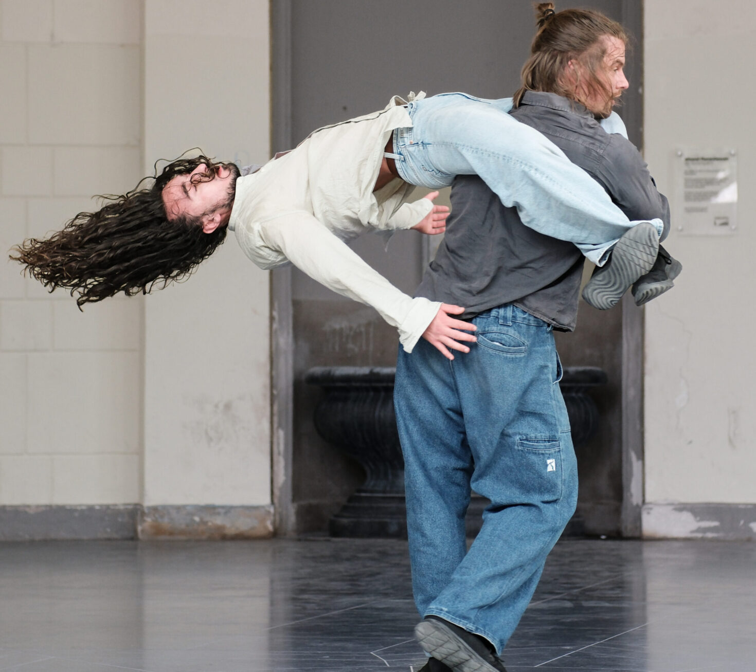 Irene K. - Two people perform a zeitgenössischer Tanz move indoors; one lifts the other horizontally in the air, long hair and arms extended back, both in casual jeans and shirts—a dynamic scene echoing Eupen's creative spirit.