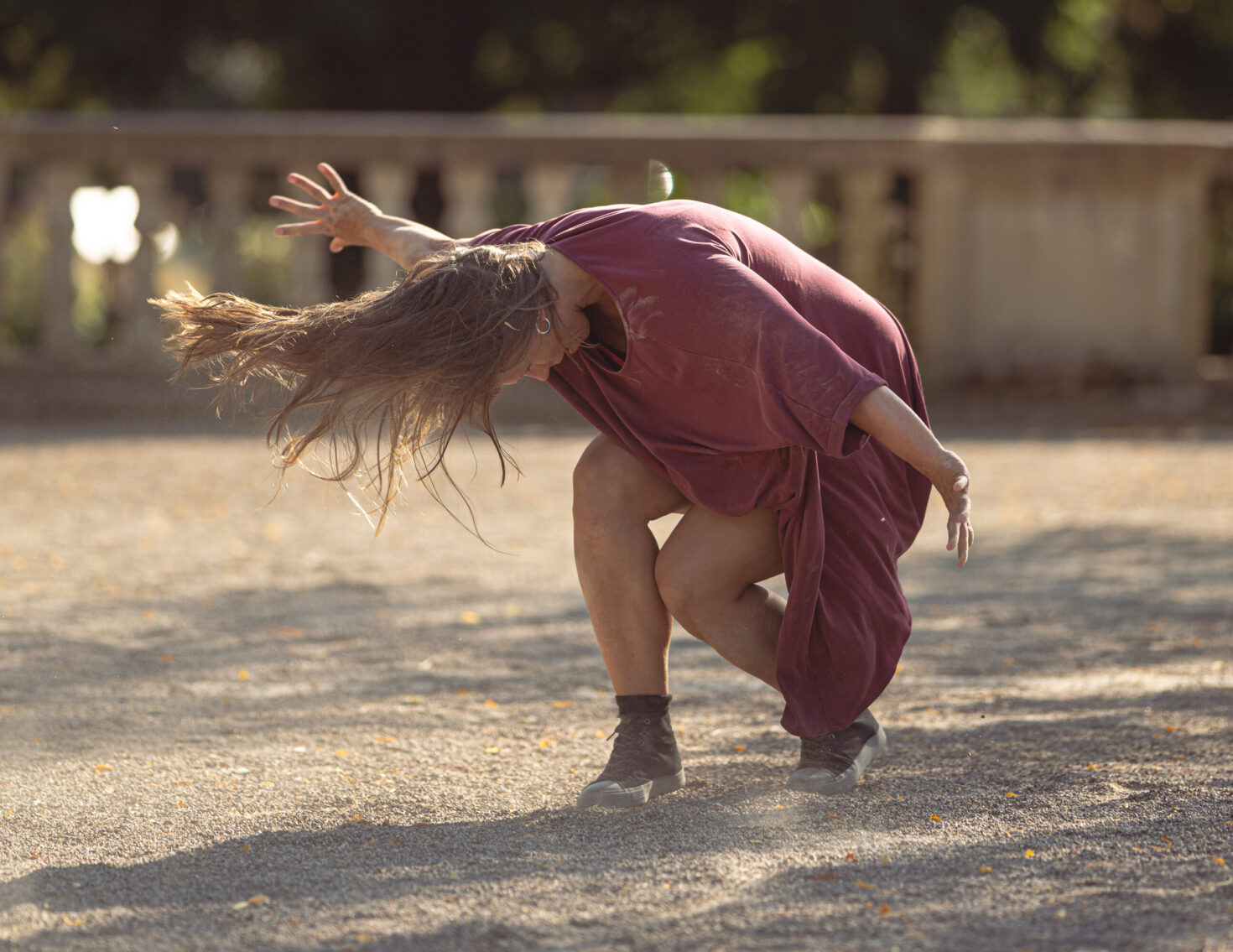Irene K. - Eine Person mit langen Haaren, die ein lockeres lila Kleid und Stiefel trägt, beugt sich an einem sonnigen Tag im Freien in einer dramatischen Tanzpose tief zum Boden hin.