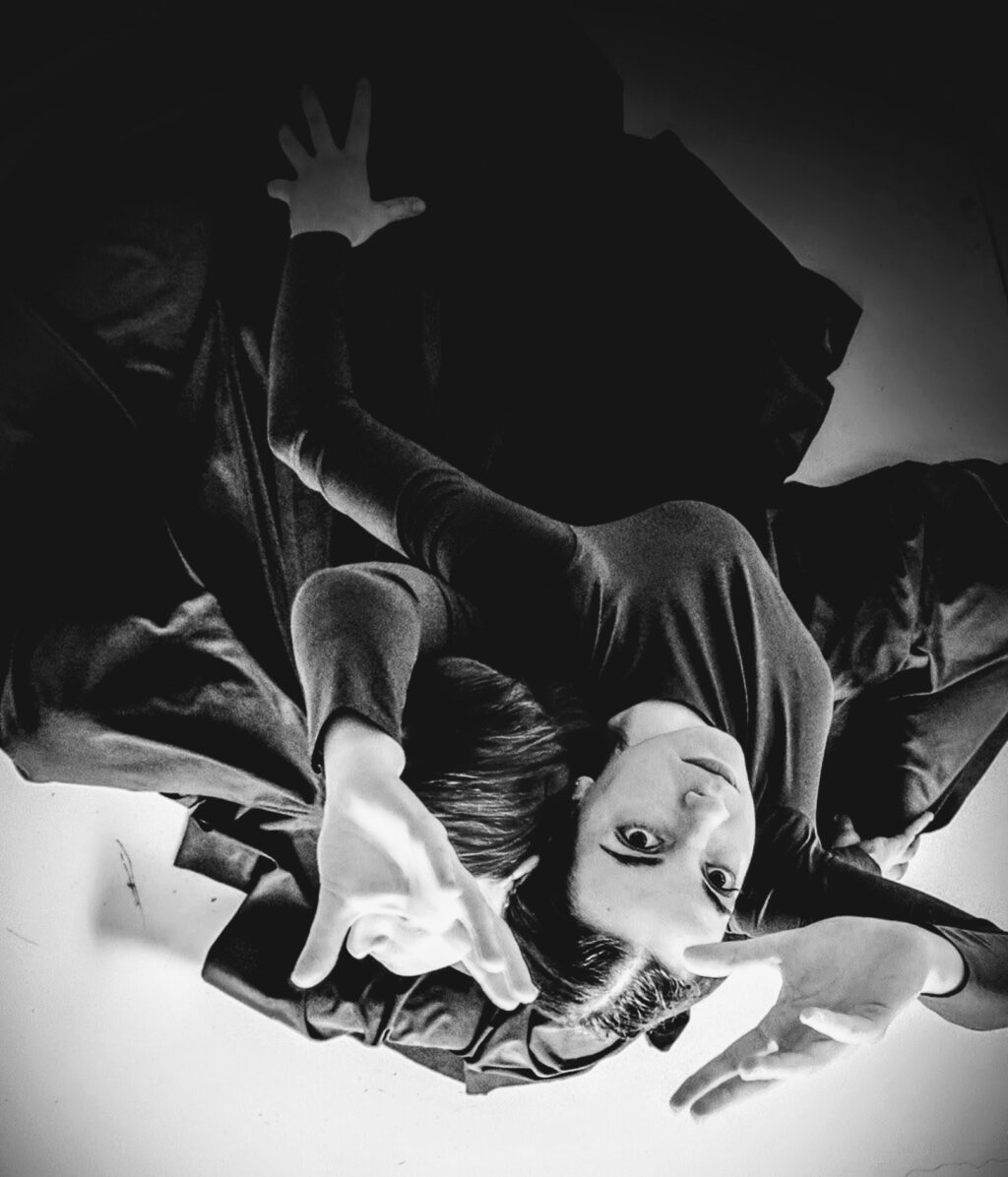 Irene K. - Une personne vêtue de noir est allongée sur une surface claire, le regard tourné vers le haut, les bras pliés et les mains écartées, créant ainsi une pose expressive et abstraite. La photo en noir et blanc renforce l'effet dramatique et artistique.