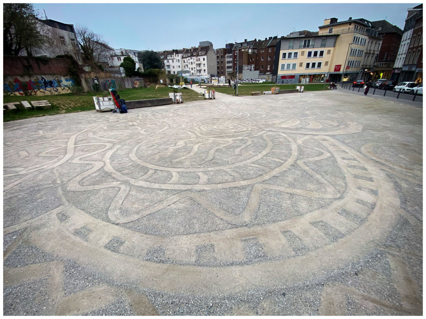 Irene K. - Auf einem sandfarbenen Stadtplatz ist ein großes, kreisförmiges Sonnenmotiv kunstvoll gezeichnet; umgeben von Bänken, Gebäuden, Graffiti und einigen wenigen Menschen an einem bewölkten Tag.
