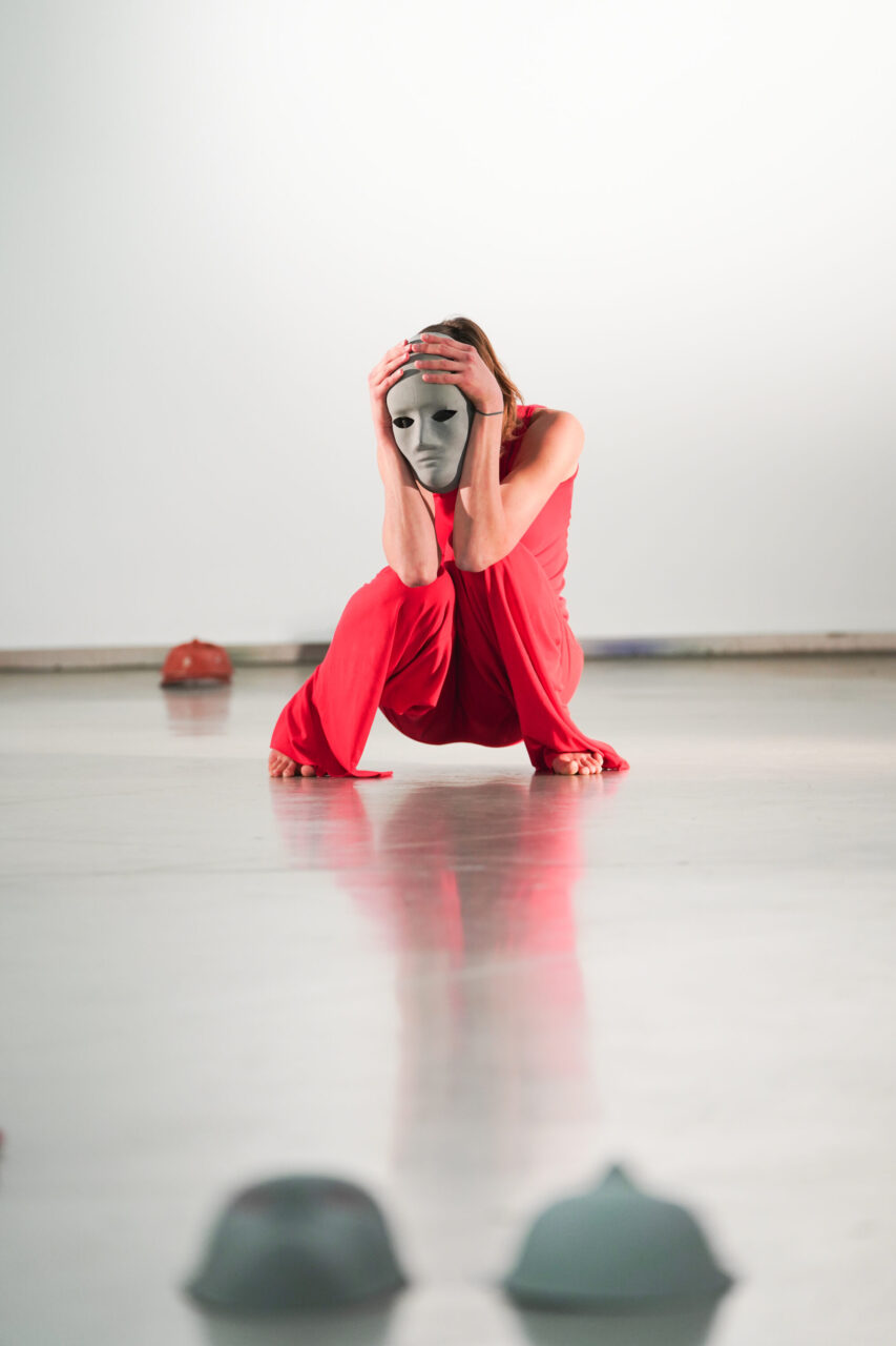 Irene K. - Eine Person in einem roten Outfit hockt auf einem reflektierenden Boden und hält sich mit beiden Händen eine weiße Maske vor das Gesicht – eine eindrucksvolle Szene aus einer Tanzperformance der Compagnie Irene K, in der im Hintergrund und im Vordergrund weitere Masken verstreut liegen.