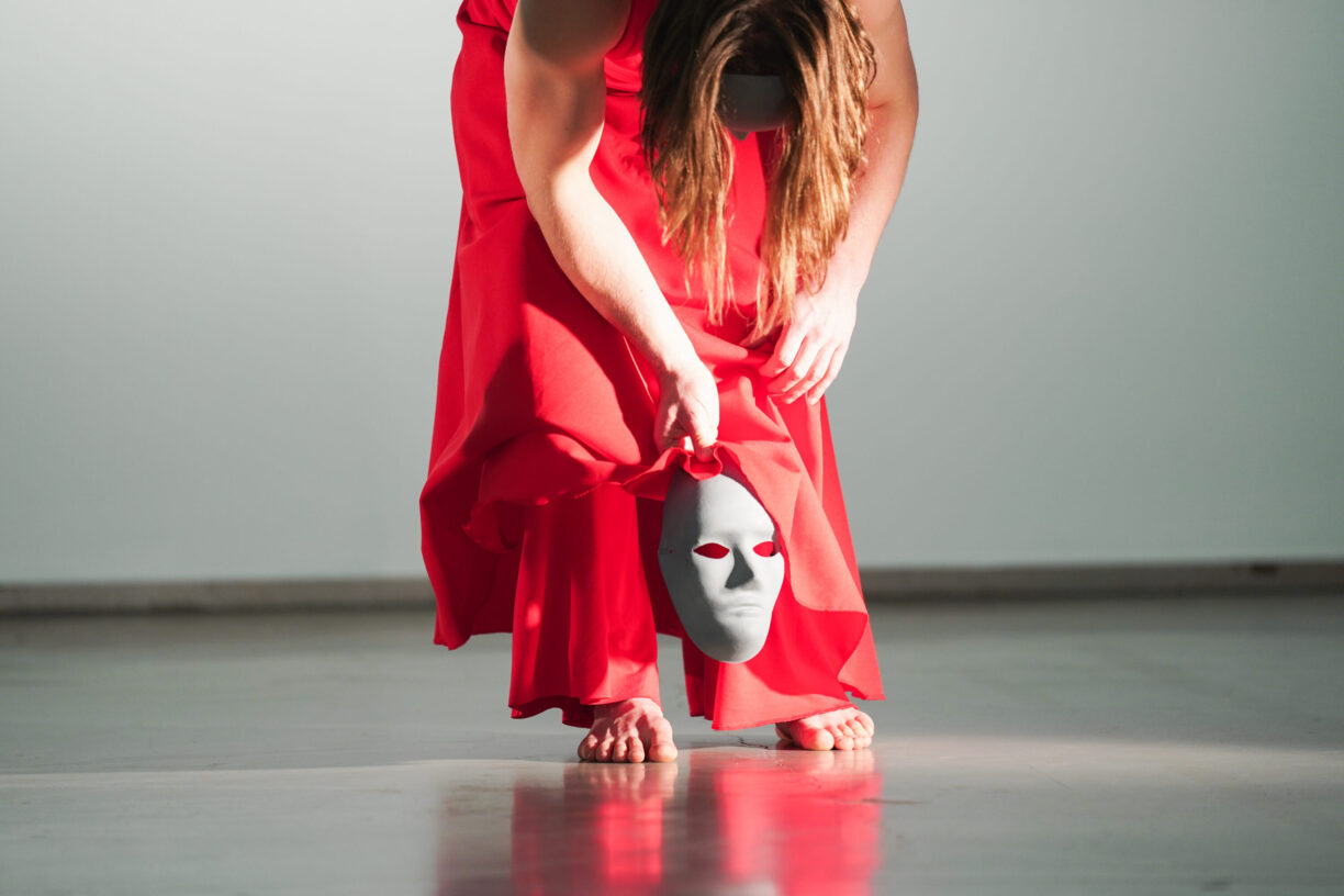 Irene K. - Eine Person in einem fließenden roten Kleid bückt sich und hält eine weiße Maske mit roten Augenlöchern. Ihr Kopf ist gesenkt und ihre nackten Füße stehen auf einem glatten Boden - eine stimmungsvolle, vom zeitgenössischen Tanz inspirierte Szene mit sanfter Beleuchtung im Hintergrund.