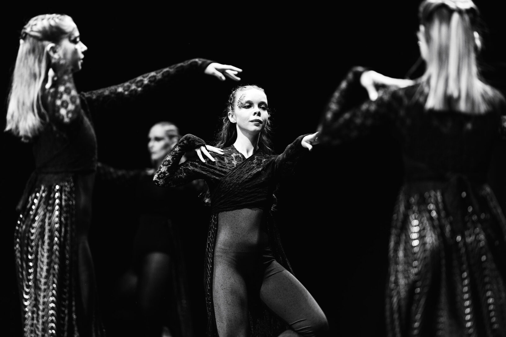 Irene K. - Vier Tänzerinnen in dunklen, gemusterten Kostümen treten auf der Bühne in einer dramatischen Schwarz-Weiß-Tanzaufführung auf. Die Tänzerin in der Mitte blickt mit eindringlichem Gesichtsausdruck nach vorne, während die anderen um sie herum posieren und ihre Arme in einem kreativen Tanz erhoben halten.