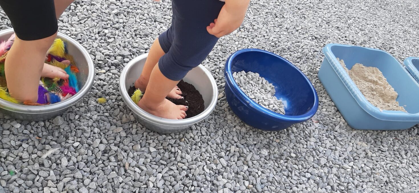 Irene K. - Ein Kind läuft barfuß durch verschiedene Sinnesschalen, die mit Federn, Erde, Steinen und Sand gefüllt sind – wie ein spielerischer Tanzkurs für die Sinne –, die alle auf einer Kiesfläche stehen. Nur die Beine und Füße des Kindes sind zu sehen.