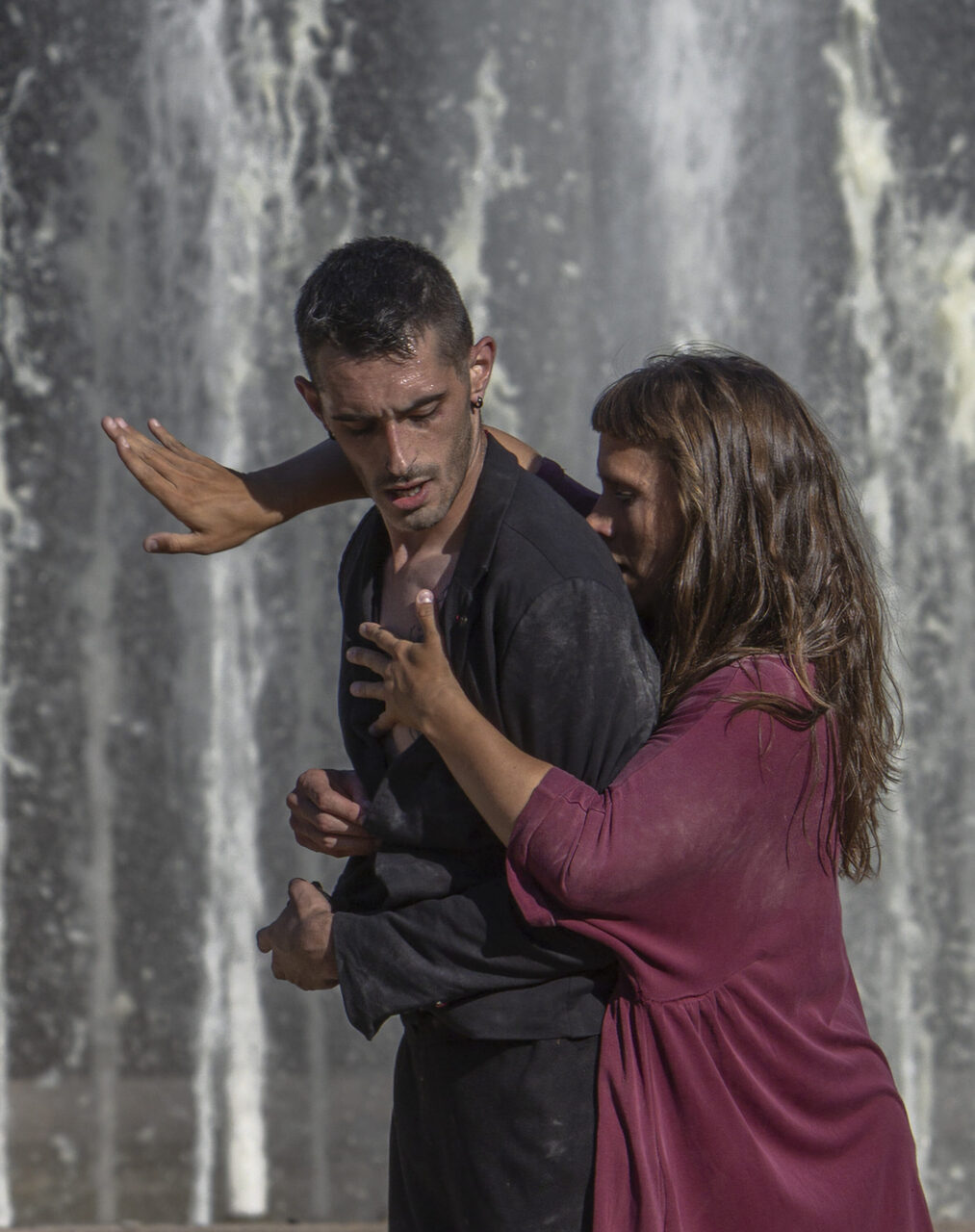 Irene K. - Deux personnes exécutent une danse contemporaine devant une fontaine en cascade. L'homme porte des vêtements sombres, tandis que la femme, vêtue d'une robe bordeaux, lui touche l'épaule et la poitrine ; tous deux arborent des expressions intenses.
