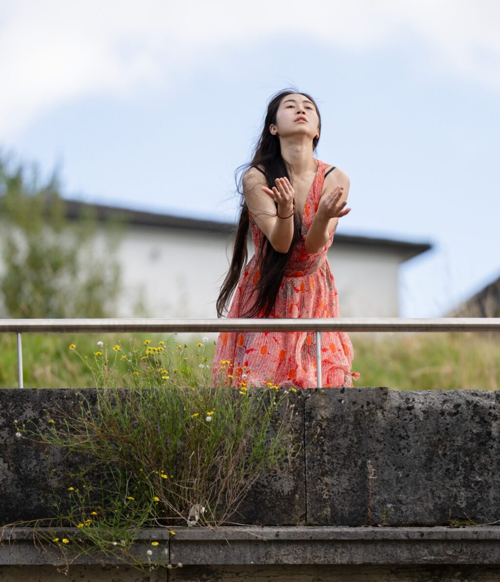 Irene K. - Eine Frau in einem korallenfarbenen Kleid steht hinter einer Steinmauer mit grünen Pflanzen, hebt die Arme und blickt nach oben, im Hintergrund sind ein Gebäude und der blaue Himmel zu sehen.