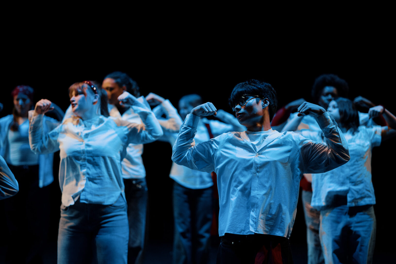 Irene K. - Eine Gruppe von Menschen in weißen Hemden führt auf der Bühne des Tanzstudios Eupen eine synchronisierte Tanzchoreografie auf, wobei jeder die Arme angewinkelt und die Fäuste nahe an den Schultern hält, beleuchtet von dramatischer Beleuchtung.